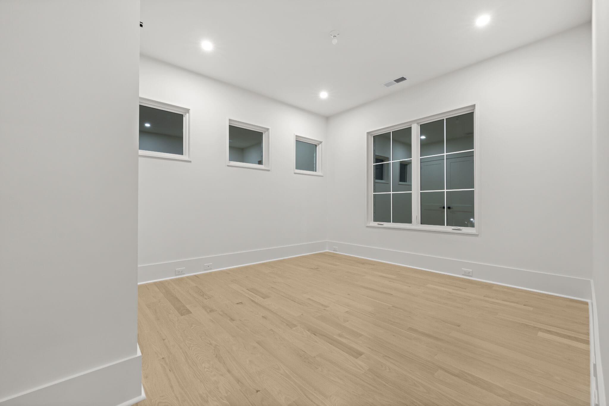 Empty room with light-colored wooden flooring, white walls, and five windows: three small windows on one wall and two larger windows on an adjacent wall. Ceiling lights are on.