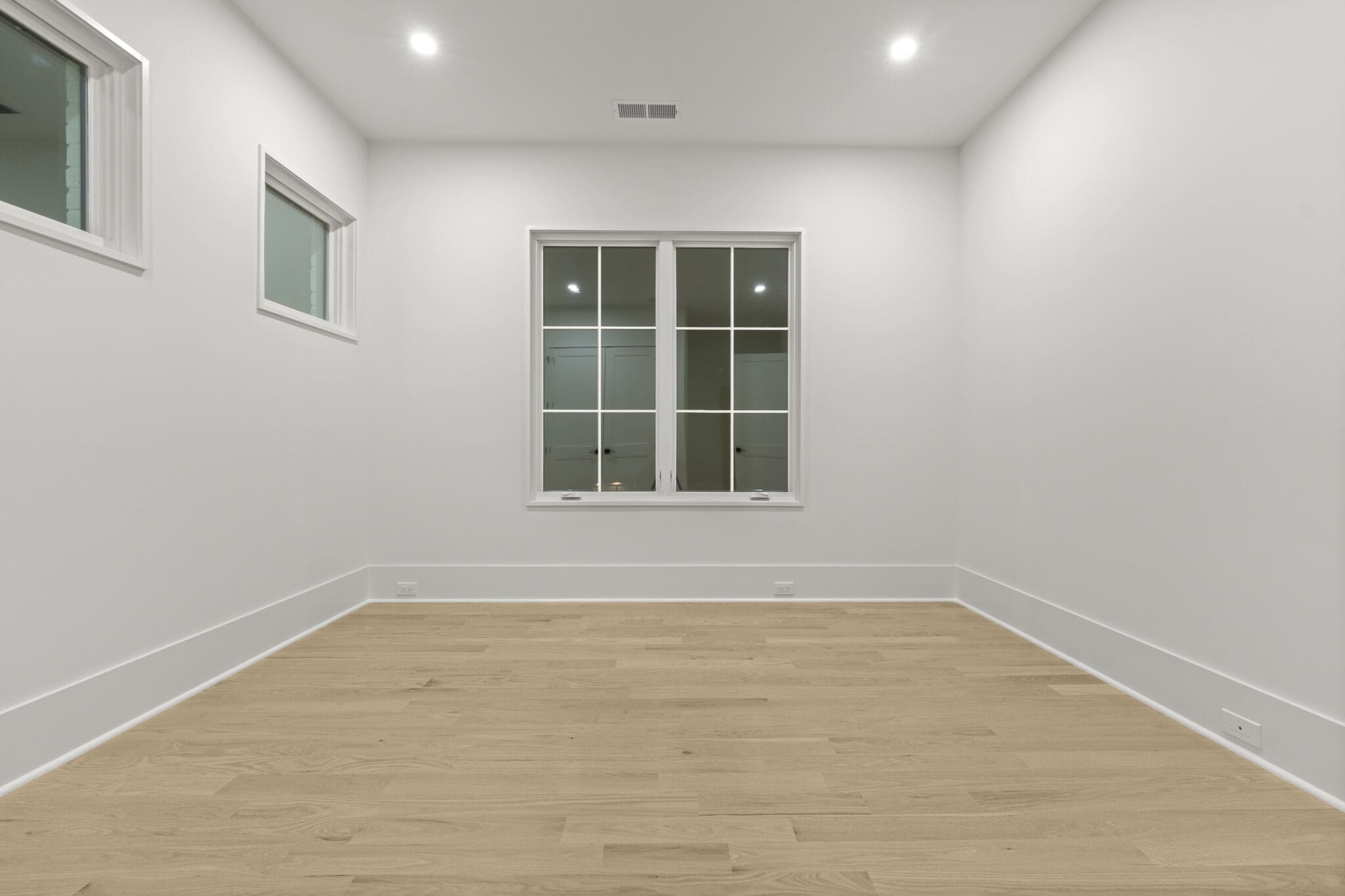 A vacant room with white walls, light wooden flooring, two small windows on the left, and a large windowed door at the far end.