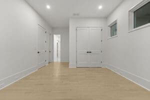 A modern, unfurnished room with wooden flooring, white walls, two closed double doors, two small windows, and recessed ceiling lights. A hallway is visible in the background.
