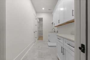 A modern laundry room with white cabinets, a washer and dryer, a sink, and a countertop. The floor is tiled, and a walk-in closet is visible in the background.