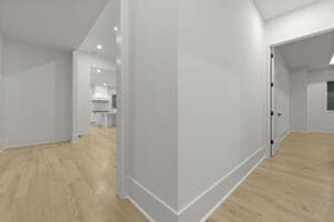 A hallway in a modern home with light wooden floors and white walls. The view includes an adjacent room with large windows and a partially visible kitchen with white cabinetry and a range hood.