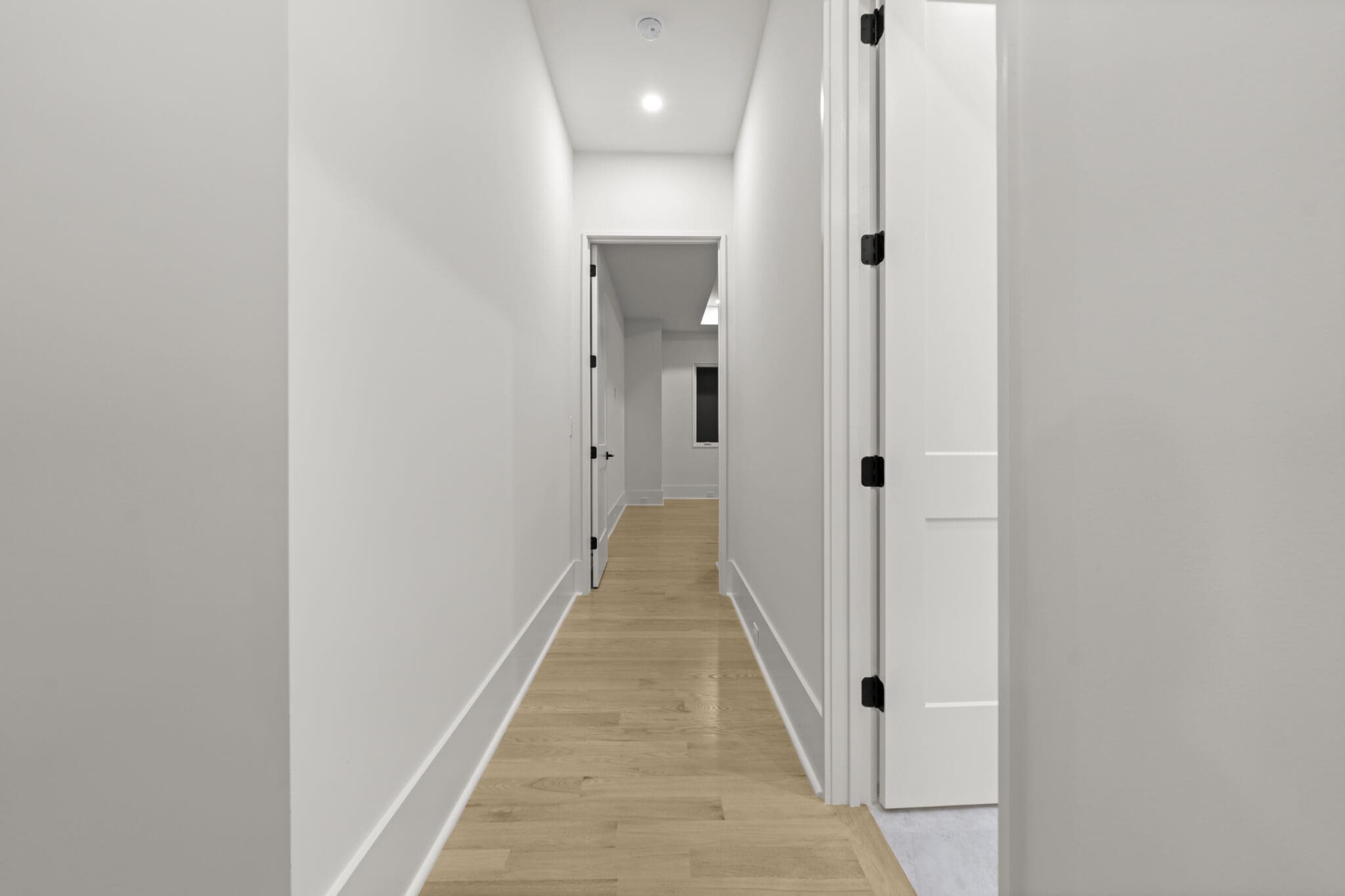 A long hallway with light wooden flooring, white walls, and several doors on both sides, leading to a room at the end.