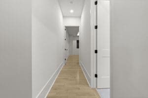 A long hallway with light wooden flooring, white walls, and several doors on both sides, leading to a room at the end.