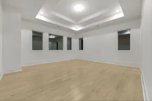 A spacious empty room with light-colored hardwood floors, white walls, five windows, and a ceiling with recessed lighting.
