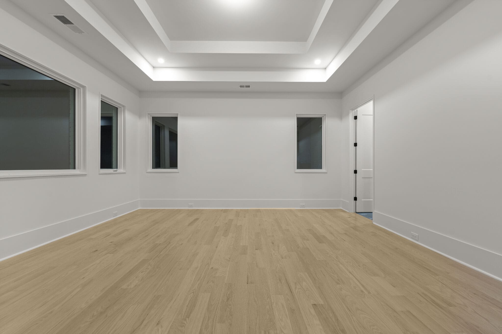 A spacious, unfurnished room with light wooden floors, white walls, a tray ceiling, and several windows. There is a closed door to the right side of the room.