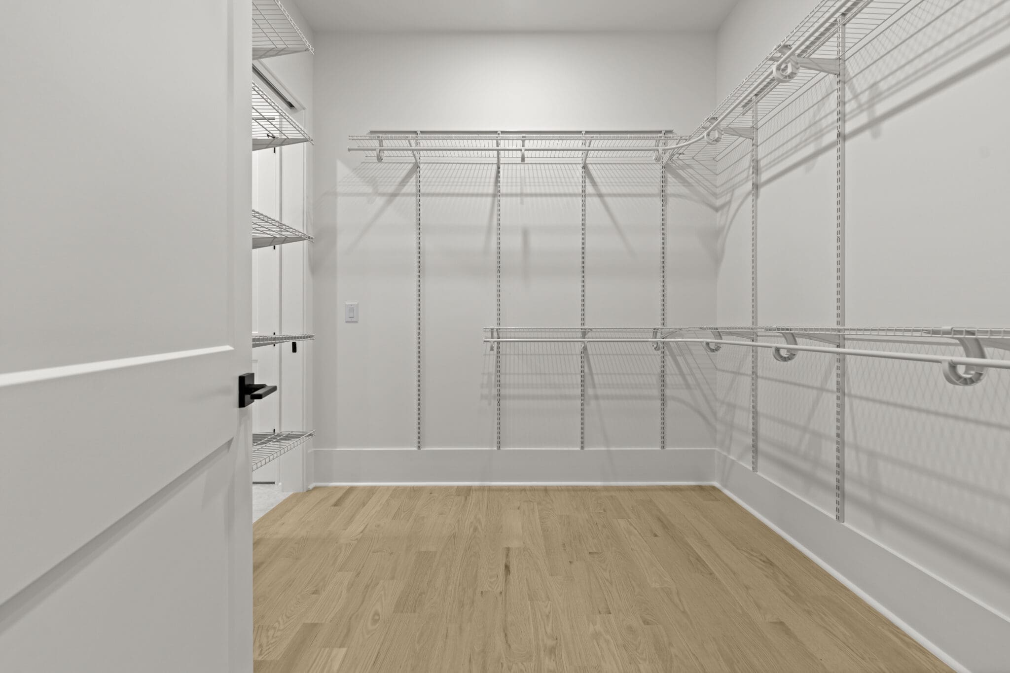 A spacious, empty walk-in closet with white wire shelving and light wood flooring. The door is partially open on the left side of the image.