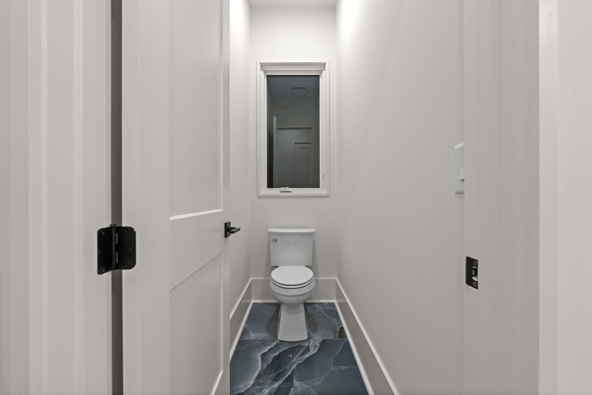 A small, white-walled bathroom features a toilet and a single window. The floor is covered with dark, marble-patterned tiles. A closed door is on the left side.