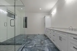 Modern bathroom with glass-enclosed shower, white vanity with multiple drawers, double sinks, and blue-gray patterned floor tiles. White walls and ceiling with recessed and wall-mounted lighting.