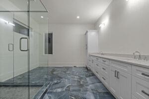 Modern bathroom with a large glass-enclosed shower, blue marbled floor tiles, long white vanity with double sinks, and white walls.