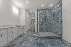A modern bathroom features white cabinetry with multiple drawers, a beige countertop, and a large glass-enclosed shower. The floor and shower walls are tiled with blue and gray marble.