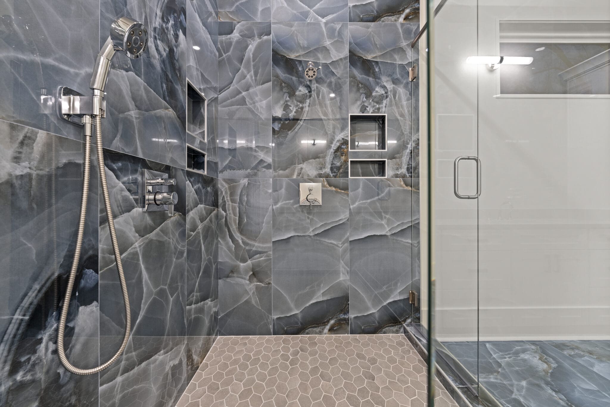 Modern shower with grey and blue marbled tiles, metal shower-head, hexagon floor tiles, and glass door. Two built-in shelves, one with a niche containing a decorative item, are visible on the wall.