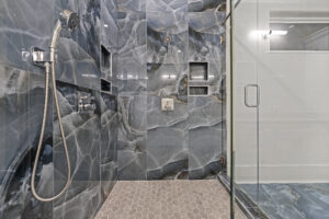 Modern shower with grey and blue marbled tiles, metal shower-head, hexagon floor tiles, and glass door. Two built-in shelves, one with a niche containing a decorative item, are visible on the wall.