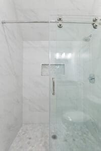 A modern shower with white tile walls, a glass door, and a chrome handle. It features a hexagonal tile floor, a wall niche for toiletries, and a ceiling-mounted showerhead.