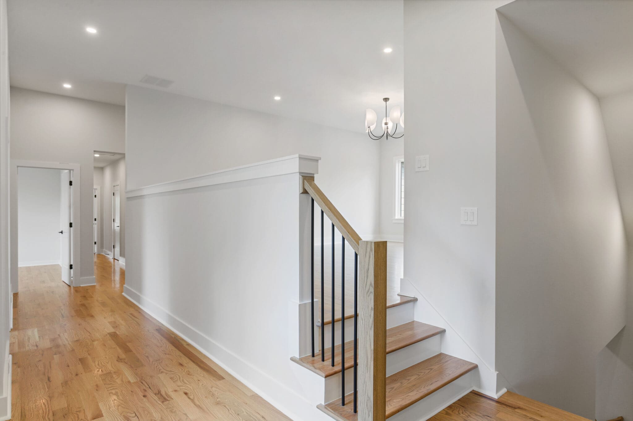 Modern hallway with light wood flooring, white walls, a staircase with black railings, and a chandelier. Doorways are visible at the end of the hallway.