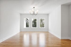 A spacious, empty room with hardwood flooring, white walls, a modern chandelier, and three large windows overlooking trees.